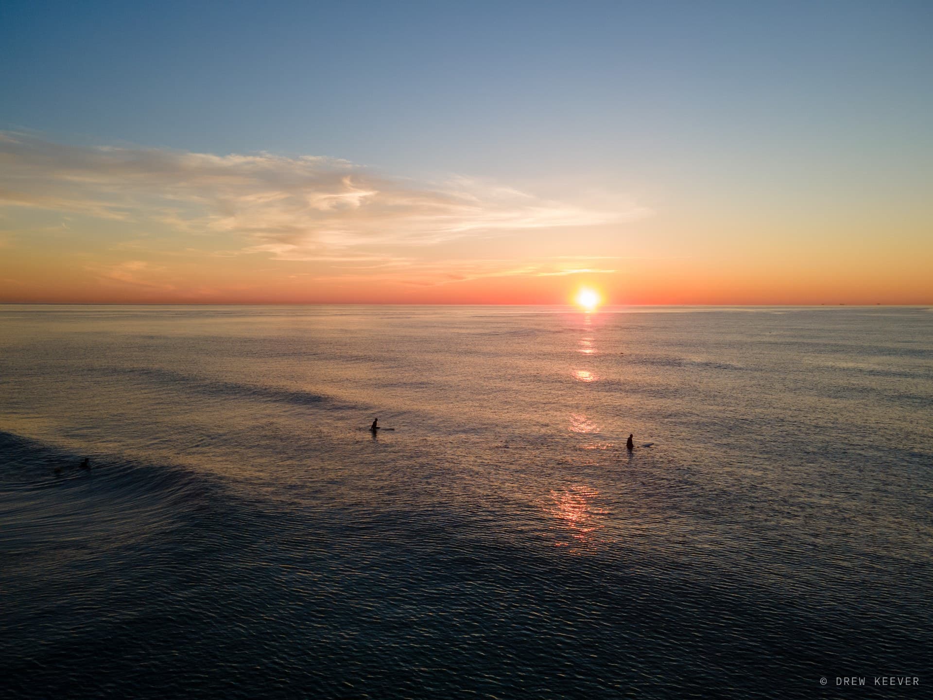 Aerial view of surfers at first light off Virginia Beach — two silhouettes waiting on a glassy Atlantic as the sun clears the horizon