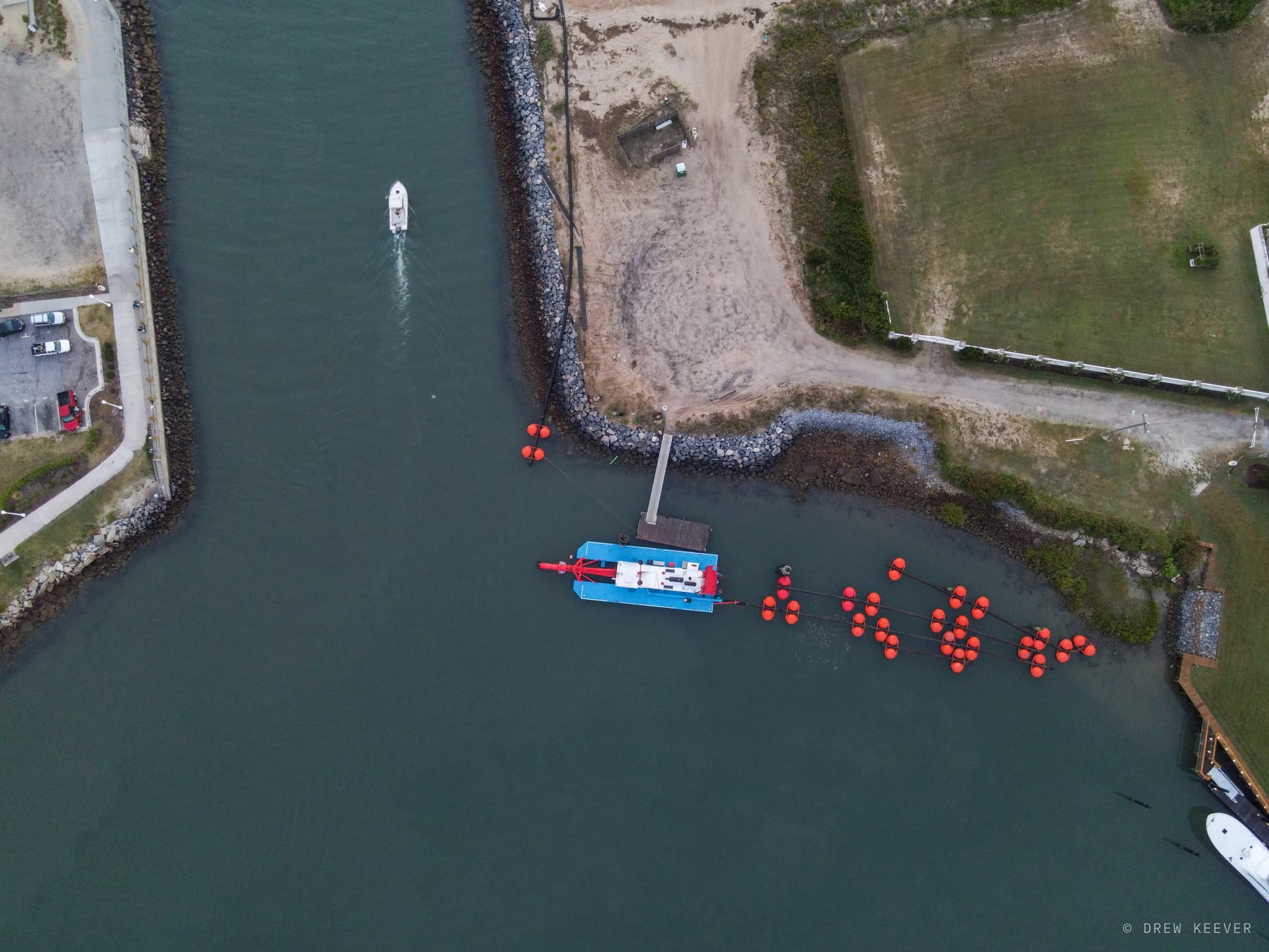 Top-down aerial of a blue dredge boat tending a line of orange buoys at the mouth of Rudee Inlet while a boat runs up the channel