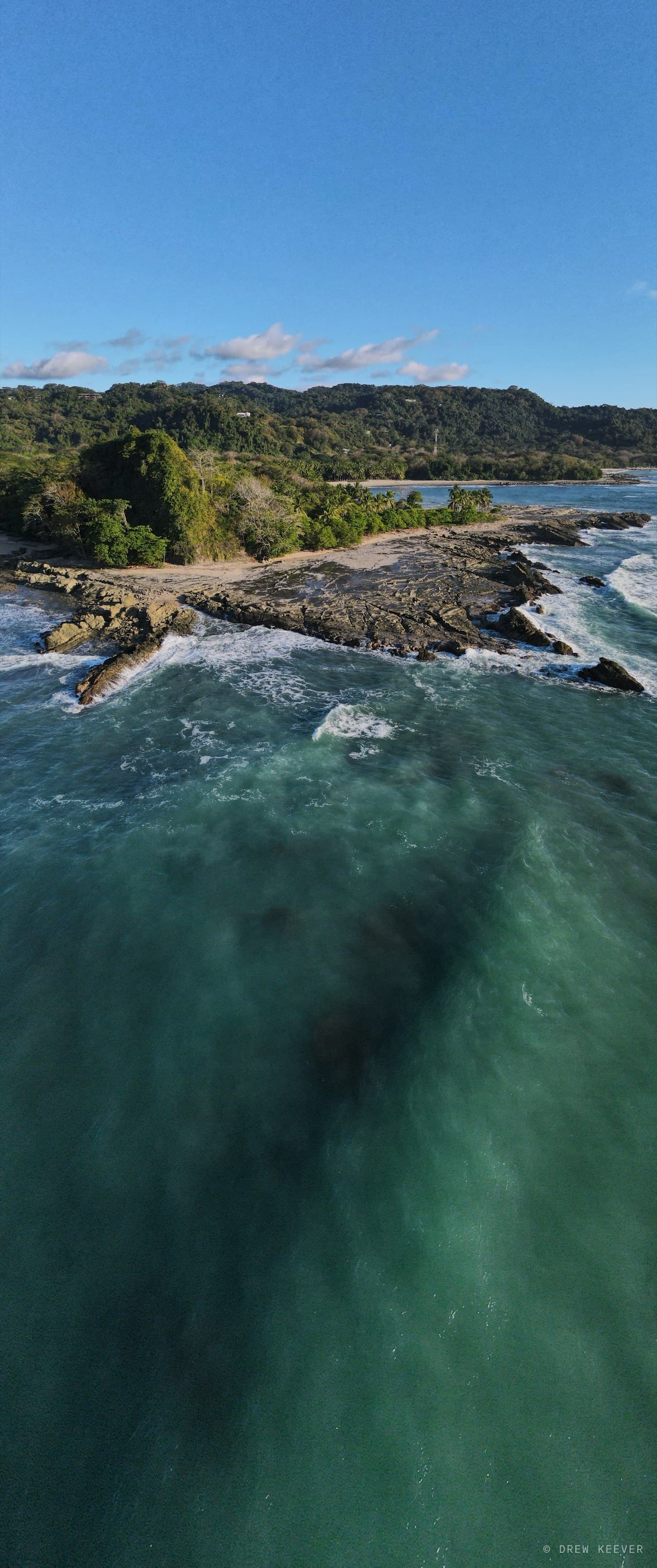 Vertical aerial panorama over the rocky point near Santa Teresa, Costa Rica — reef shadow and jade Pacific on the Nicoya Peninsula