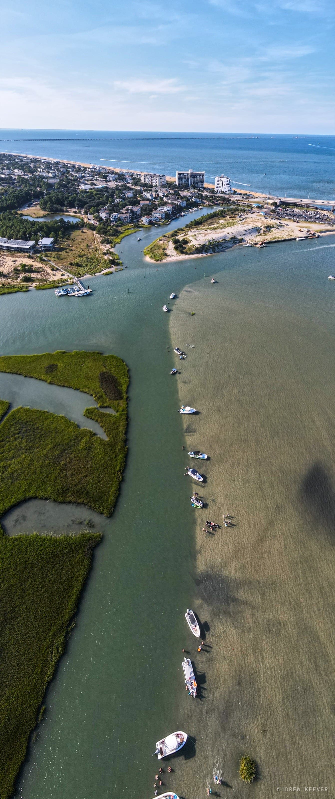 Tall aerial of boats anchored in a line along the edge of the Lynnhaven Inlet sandbar on a summer Saturday, swimmers in the shallows and the Chesapeake Bay beyond
