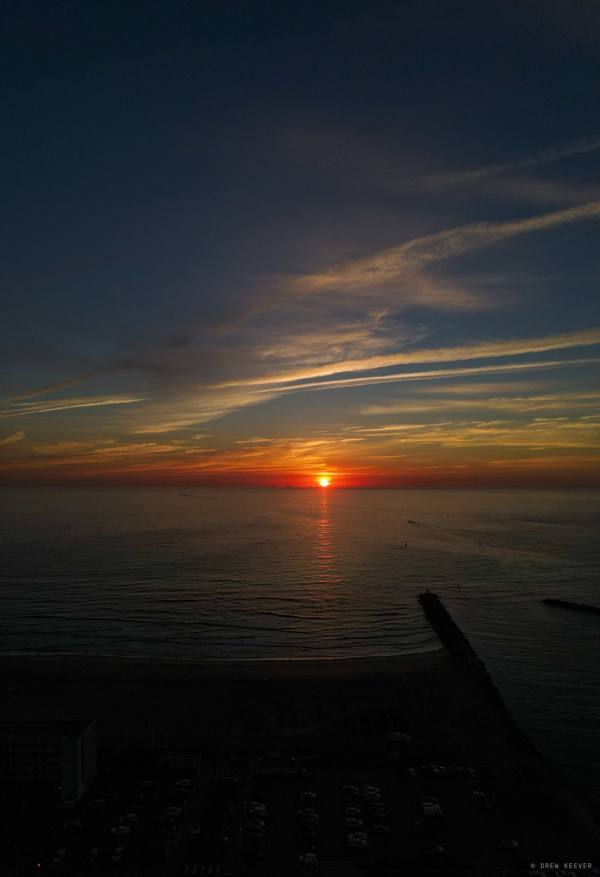 Vertical aerial at sunrise — the sun clearing the Atlantic horizon, waves breaking on a dark beach and jetty below