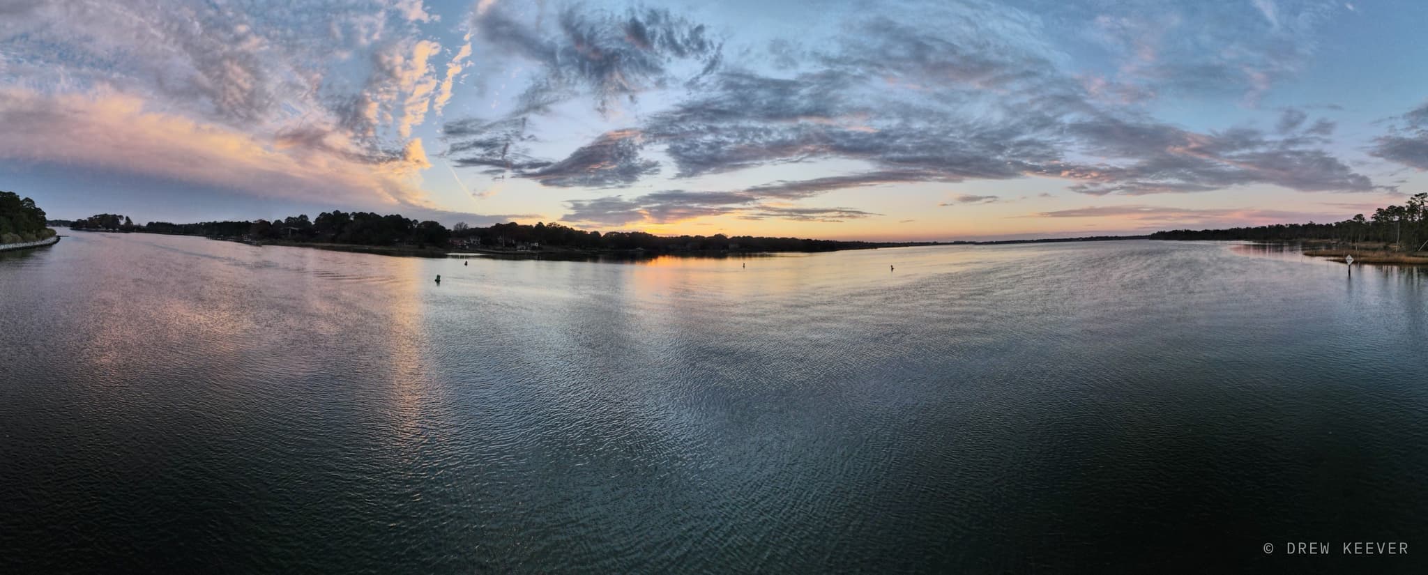 Wide panorama of a flat, calm tidal river at sunset, the sky's pinks and golds reflected in the still water