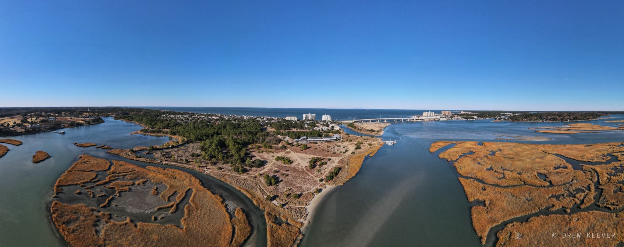 Aerial winter panorama of golden salt marsh and winding tidal channels, with the Lesner Bridge and the bay on the horizon
