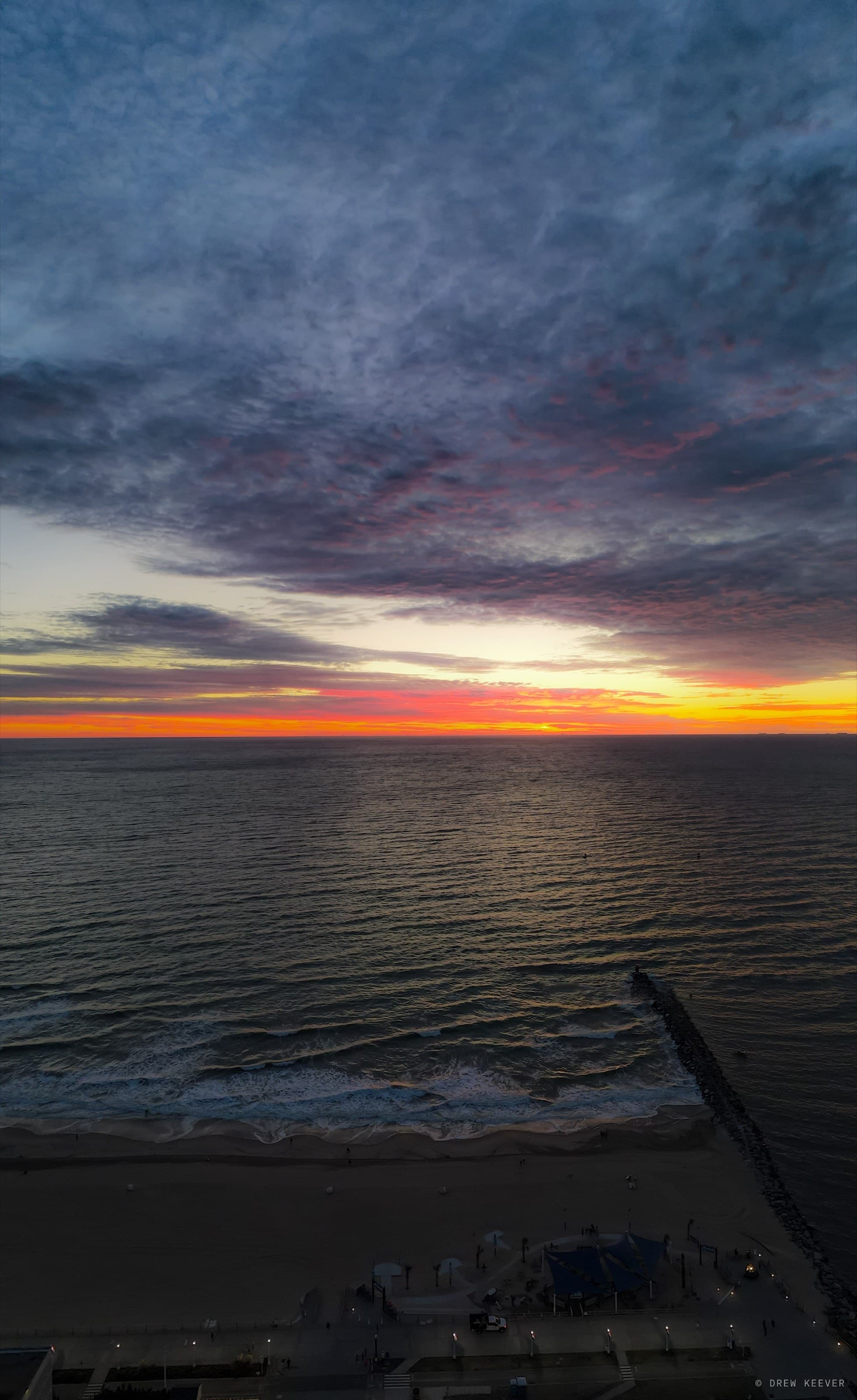 Vertical aerial at dusk — violet and pink clouds over the Atlantic, waves washing the beach beside the Virginia Beach boardwalk