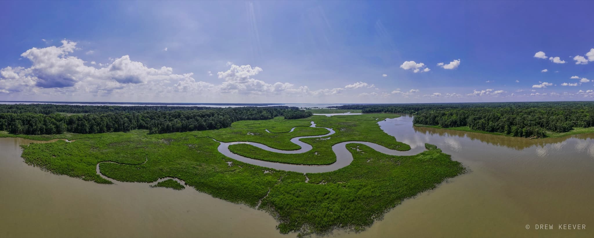Aerial panorama of a tidal creek meandering in tight S-curves through bright green salt marsh near Williamsburg, Virginia, with a wide brown river beyond